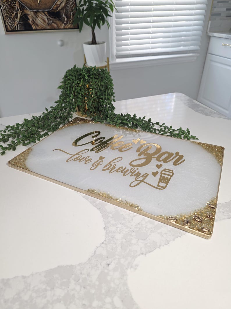 May include: A rectangular coffee bar serving tray with a white and gold design. The tray has gold glitter accents on the edges and corners, with the words "Coffee Bar love is brewing" in gold script. A coffee cup icon is also included.