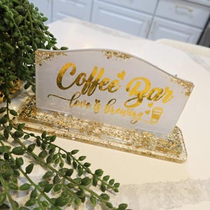 K&ouml;nnte beinhalten: Ein rechteckiges Schild aus wei&szlig;em Harz mit Blattgoldflocken und dem goldenen Schriftzug Coffee Bar love is brewing mit kleinen Herzsymbolen und einer Kaffeetassengrafik. Das Schild ist auf einem passenden rechteckigen Harzsockel montiert.