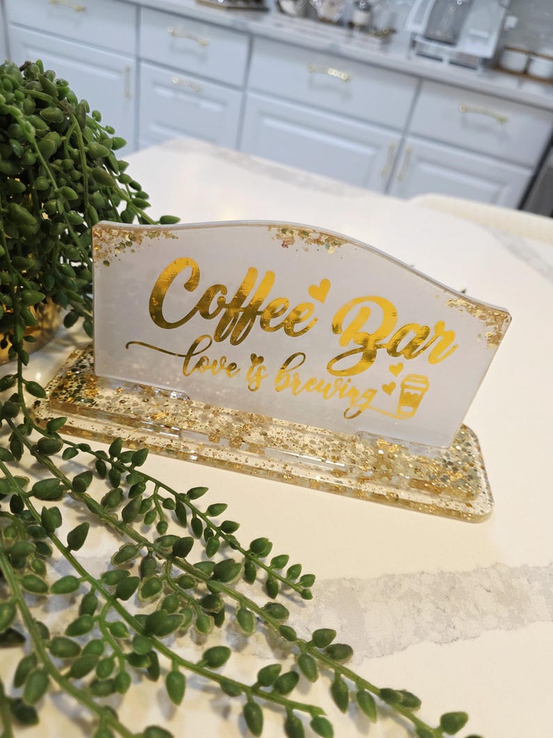 K&ouml;nnte beinhalten: Ein rechteckiges Schild aus wei&szlig;em Harz mit Blattgoldflocken und dem goldenen Schriftzug Coffee Bar love is brewing mit kleinen Herzsymbolen und einer Kaffeetassengrafik. Das Schild ist auf einem passenden rechteckigen Harzsockel montiert.