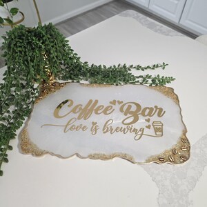 May include: A white and gold coffee bar sign with the words "Coffee Bar" and "love is brewing" in gold lettering. The sign has a gold glitter border and a coffee cup graphic.
