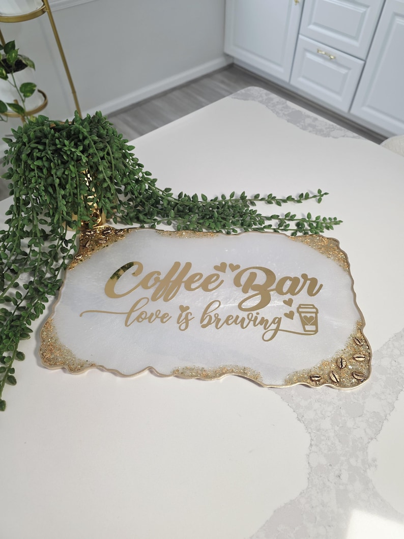 May include: A white and gold coffee bar sign with the words "Coffee Bar" and "love is brewing" in gold lettering. The sign has a gold glitter border and a coffee cup graphic.