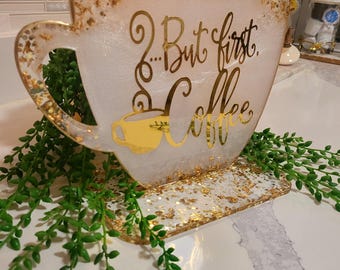 May include: A decorative coffee cup-shaped sign with the text "...But first, Coffee" in gold script. The sign is white with gold glitter accents and a gold coffee cup illustration. Green foliage surrounds the base.