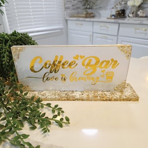 K&ouml;nnte beinhalten: Ein rechteckiges Schild aus wei&szlig;em Harz mit Blattgoldakzenten und einem goldenen St&auml;nder. Das Schild tr&auml;gt den Text Coffee Bar und love is brewing in goldener Schreibschrift, erg&auml;nzt durch kleine Herzsymbole und eine goldene Kaffeetassengrafik.