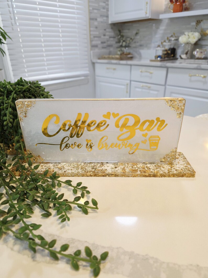 K&ouml;nnte beinhalten: Ein rechteckiges Schild aus wei&szlig;em Harz mit Blattgoldakzenten und einem goldenen St&auml;nder. Das Schild tr&auml;gt den Text Coffee Bar und love is brewing in goldener Schreibschrift, erg&auml;nzt durch kleine Herzsymbole und eine goldene Kaffeetassengrafik.