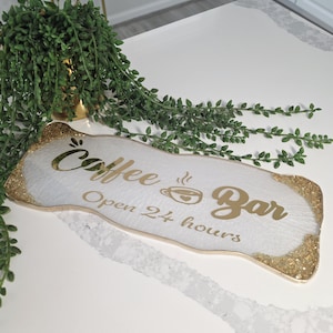 May include: A white Coffee Bar sign with gold glitter trim and gold lettering. The sign reads "Coffee Bar Open 24 hours" with a coffee cup icon. Green leafy vines and a gold pot are in the background.