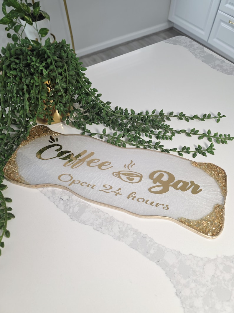 May include: A white Coffee Bar sign with gold glitter trim and gold lettering. The sign reads "Coffee Bar Open 24 hours" with a coffee cup icon. Green leafy vines and a gold pot are in the background.