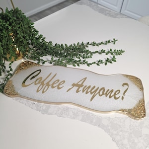 May include: A white rectangular tray with gold glitter edges and gold text that reads "Coffee Anyone?". The tray is on a white surface, with a green plant in the background.