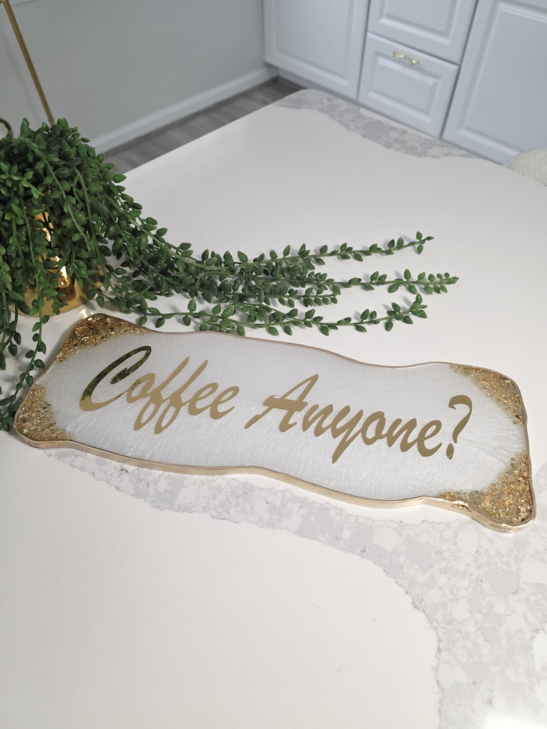 May include: A white rectangular tray with gold glitter edges and gold text that reads "Coffee Anyone?". The tray is on a white surface, with a green plant in the background.