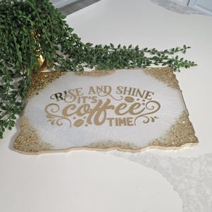 May include: A rectangular white tray with gold glitter edges and gold lettering that reads "Rise and Shine It's Coffee Time". The tray is on a white surface with a green plant in the background.