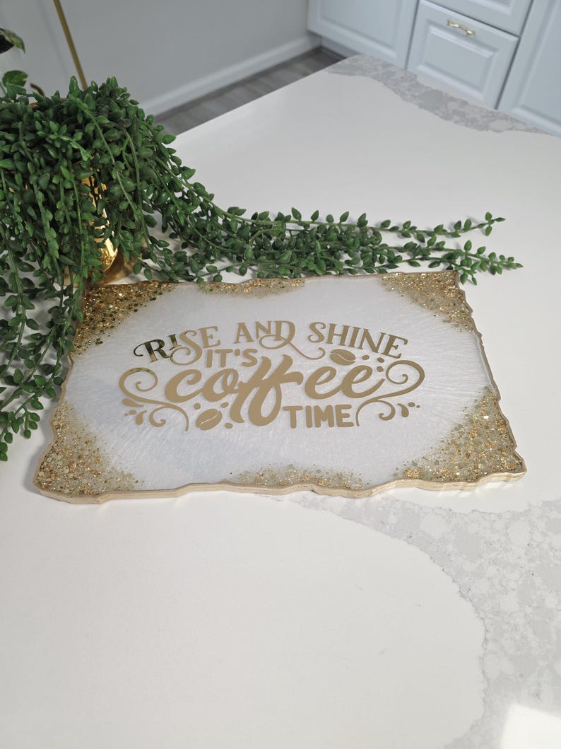 May include: A rectangular white tray with gold glitter edges and gold lettering that reads "Rise and Shine It's Coffee Time". The tray is on a white surface with a green plant in the background.