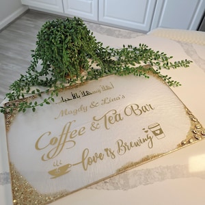 May include: A rectangular white sign with gold lettering that reads "Coffee & Tea Bar" and "love is brewing." The sign has a gold glitter border and a small coffee cup graphic. A green plant sits above the sign.