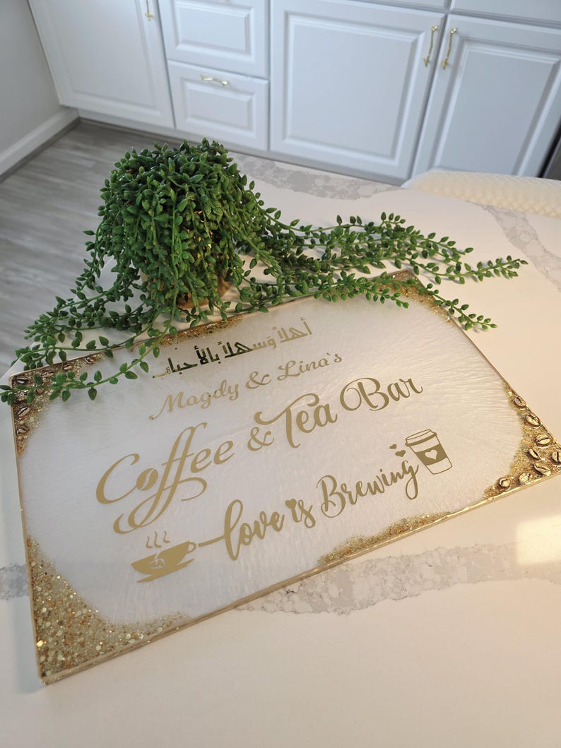 May include: A rectangular white sign with gold lettering that reads "Coffee & Tea Bar" and "love is brewing." The sign has a gold glitter border and a small coffee cup graphic. A green plant sits above the sign.