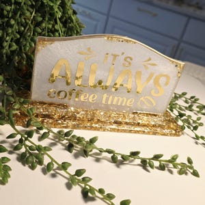 K&ouml;nnte beinhalten: Ein rechteckiges Schild aus Kunstharz mit einer wei&szlig;en Basis mit Goldglitter und einem goldfarbenen St&auml;nder. Das Schild tr&auml;gt den goldenen Schriftzug IT'S ALWAYS coffee time sowie kleine Pfeil- und Kaffeebohnensymbole.