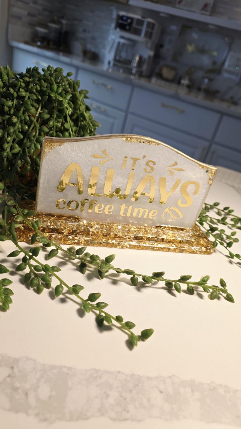 K&ouml;nnte beinhalten: Ein rechteckiges Schild aus Kunstharz mit einer wei&szlig;en Basis mit Goldglitter und einem goldfarbenen St&auml;nder. Das Schild tr&auml;gt den goldenen Schriftzug IT'S ALWAYS coffee time sowie kleine Pfeil- und Kaffeebohnensymbole.