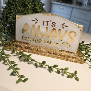 K&ouml;nnte beinhalten: Ein rechteckiges Schild aus Kunstharz mit wei&szlig;em Perlmutt-Finish und Blattgold-Flocken. Der Text "IT'S ALWAYS coffee time" ist in goldener Schrift gehalten, erg&auml;nzt durch zwei kleine Pfeile und ein Kaffeebohnen-Symbol auf einem goldenen Sockel.