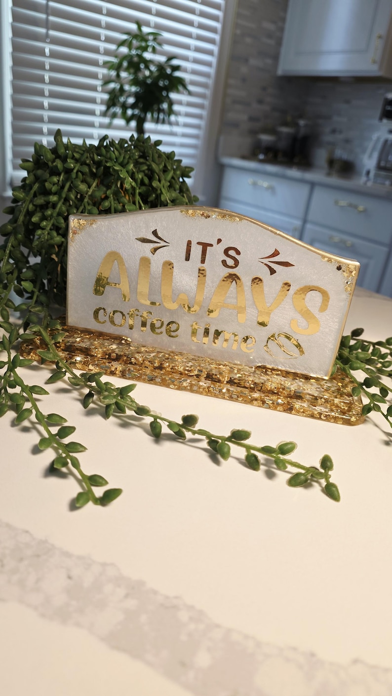 K&ouml;nnte beinhalten: Ein rechteckiges Schild aus Kunstharz mit wei&szlig;em Perlmutt-Finish und Blattgold-Flocken. Der Text "IT'S ALWAYS coffee time" ist in goldener Schrift gehalten, erg&auml;nzt durch zwei kleine Pfeile und ein Kaffeebohnen-Symbol auf einem goldenen Sockel.