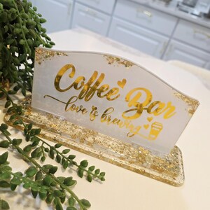 K&ouml;nnte beinhalten: Ein rechteckiges Schild aus wei&szlig;em Harz mit goldenen Glitzerakzenten und einem goldenen Sockel. Das Schild tr&auml;gt den Text "Coffee Bar" in Schreibschrift, gefolgt von "love is brewing" und einem kleinen Kaffeetassen-Symbol mit Herzen.