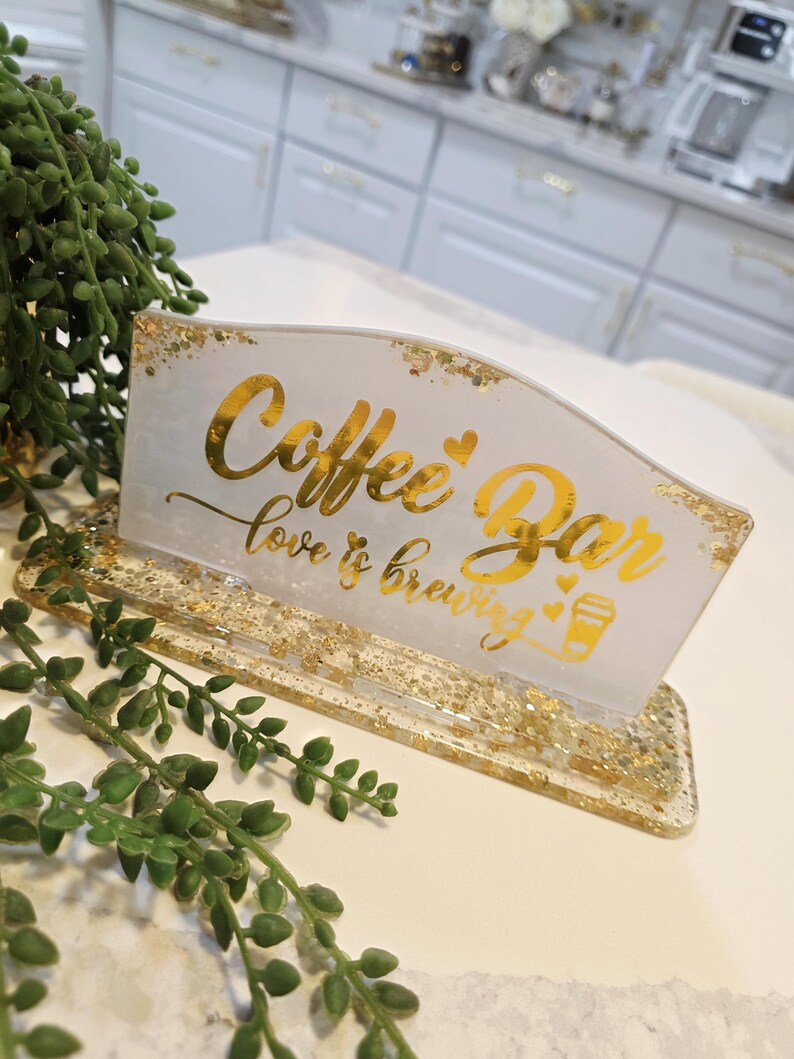 K&ouml;nnte beinhalten: Ein rechteckiges Schild aus wei&szlig;em Harz mit goldenen Glitzerakzenten und einem goldenen Sockel. Das Schild tr&auml;gt den Text "Coffee Bar" in Schreibschrift, gefolgt von "love is brewing" und einem kleinen Kaffeetassen-Symbol mit Herzen.