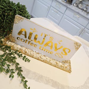 K&ouml;nnte beinhalten: Ein rechteckiges Schild aus wei&szlig;em Harz mit Blattgoldakzenten und einem goldenen St&auml;nder. Der Text auf dem Schild lautet "IT'S ALWAYS coffee time" in einer Mischung aus fetten und geschwungenen Schriftarten. Die R&auml;nder und der Sockel sind vergoldet.
