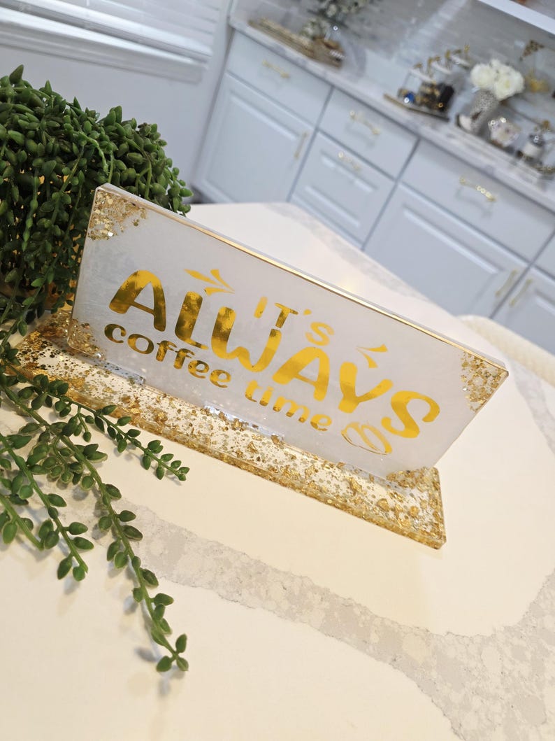 K&ouml;nnte beinhalten: Ein rechteckiges Schild aus wei&szlig;em Harz mit Blattgoldakzenten und einem goldenen St&auml;nder. Der Text auf dem Schild lautet "IT'S ALWAYS coffee time" in einer Mischung aus fetten und geschwungenen Schriftarten. Die R&auml;nder und der Sockel sind vergoldet.