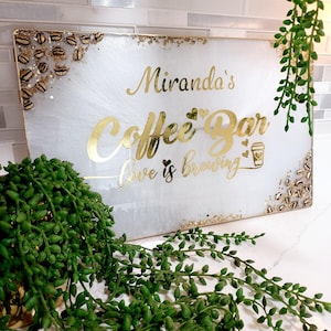 May include: A rectangular white sign with gold lettering that reads "Miranda's Coffee Bar, love is brewing." The sign has gold accents and coffee bean details. Green plants are in the foreground and background.
