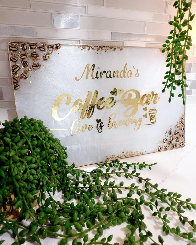 May include: A rectangular white sign with gold lettering that reads "Miranda's Coffee Bar, love is brewing." The sign has gold accents and coffee bean details. Green plants are in the foreground and background.