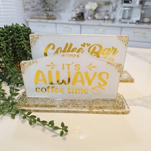K&ouml;nnte beinhalten: Zwei rechteckige wei&szlig;e Harzschilder mit Blattgoldflocken und goldener Schrift. Das hintere Schild zeigt Coffee Bar mit zwei Herzen. Das vordere Schild zeigt IT'S ALWAYS coffee time mit Pfeilen und einem Kaffeebohnensymbol. Beide Schilder haben einen Sockel.