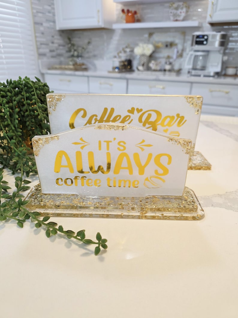 K&ouml;nnte beinhalten: Zwei rechteckige wei&szlig;e Harzschilder mit Blattgoldflocken und goldener Schrift. Das hintere Schild zeigt Coffee Bar mit zwei Herzen. Das vordere Schild zeigt IT'S ALWAYS coffee time mit Pfeilen und einem Kaffeebohnensymbol. Beide Schilder haben einen Sockel.