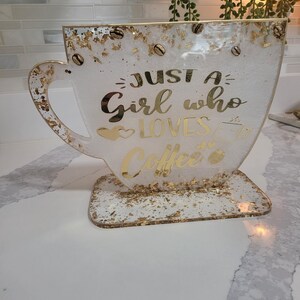 K&ouml;nnte beinhalten: Ein dekoratives Kunstwerk aus Harz in Form einer Kaffeetasse. Die wei&szlig;e Tasse hat goldene Flocken und Kaffeebohnen-Akzente. Goldener Text lautet "Just a Girl who Loves Coffee". Die Tasse steht auf einem passenden Sockel.