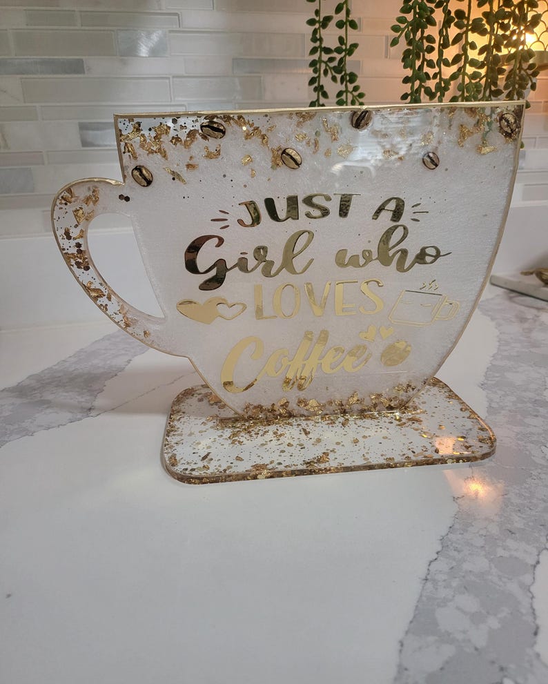 K&ouml;nnte beinhalten: Ein dekoratives Kunstwerk aus Harz in Form einer Kaffeetasse. Die wei&szlig;e Tasse hat goldene Flocken und Kaffeebohnen-Akzente. Goldener Text lautet "Just a Girl who Loves Coffee". Die Tasse steht auf einem passenden Sockel.