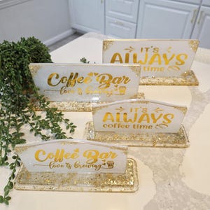 K&ouml;nnte beinhalten: Vier rechteckige Harzschilder mit goldenen Glitzerakzenten und goldener Schrift. Zwei Schilder tragen die Aufschrift Coffee Bar love is brewing und zwei IT'S ALWAYS coffee time. Jedes Schild ist auf einem passenden rechteckigen Harzsockel montiert.