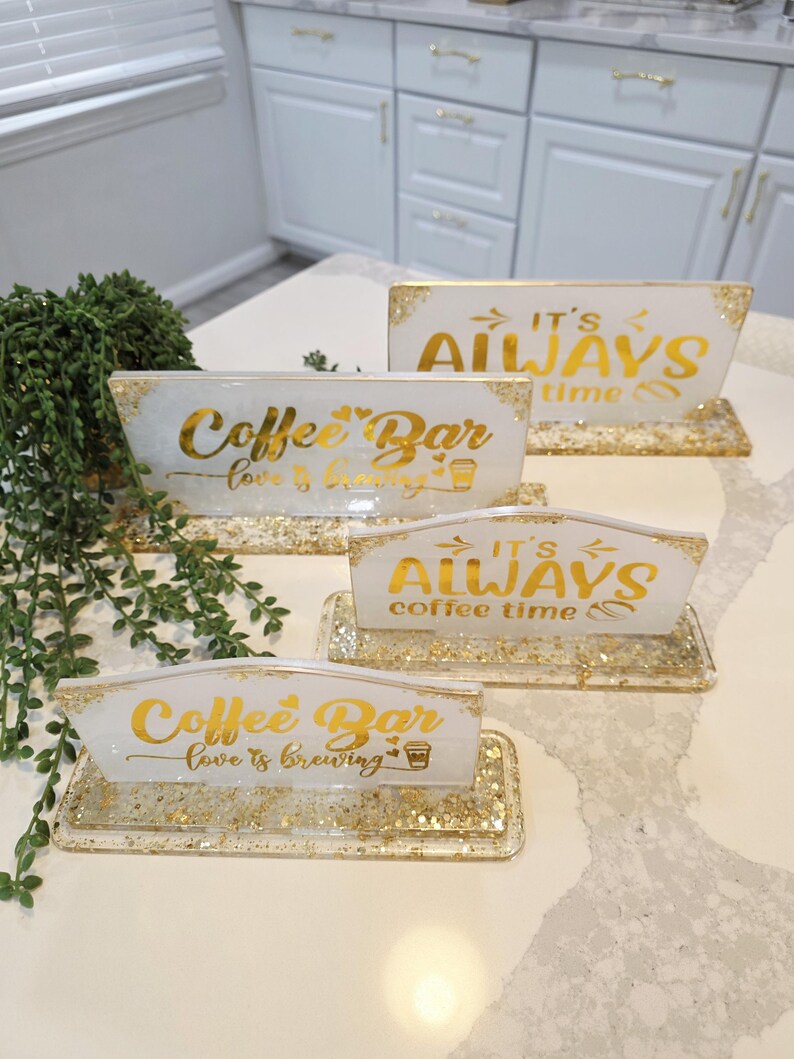K&ouml;nnte beinhalten: Vier rechteckige Harzschilder mit goldenen Glitzerakzenten und goldener Schrift. Zwei Schilder tragen die Aufschrift Coffee Bar love is brewing und zwei IT'S ALWAYS coffee time. Jedes Schild ist auf einem passenden rechteckigen Harzsockel montiert.
