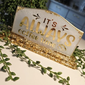 K&ouml;nnte beinhalten: Ein rechteckiges Schild aus wei&szlig;em Harz mit Blattgoldakzenten und einem goldenen Sockel. Der Text IT'S ALWAYS coffee time ist in goldener Schrift zu sehen. Das Schild hat eine glatte, gl&auml;nzende Oberfl&auml;che mit eingebetteten Goldflocken.
