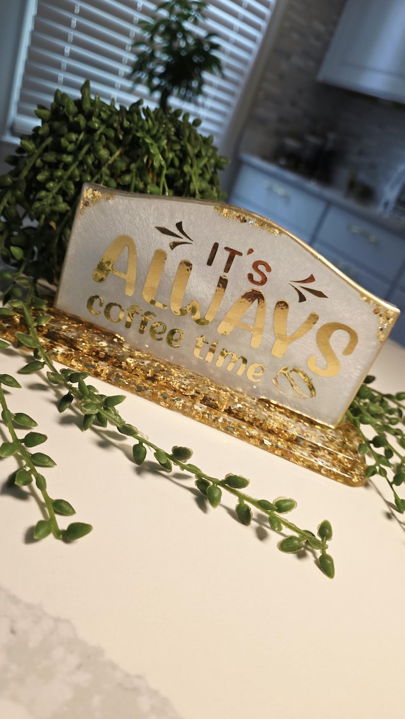 K&ouml;nnte beinhalten: Ein rechteckiges Schild aus wei&szlig;em Harz mit Blattgoldakzenten und einem goldenen Sockel. Der Text IT'S ALWAYS coffee time ist in goldener Schrift zu sehen. Das Schild hat eine glatte, gl&auml;nzende Oberfl&auml;che mit eingebetteten Goldflocken.