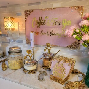 May include: A coffee and tea bar setup with a pink sign that reads "Coffee & Tea Bar" in gold lettering. The setup includes a jar, sugar bowl, and coasters, all with gold accents. A decorative lamp is visible.