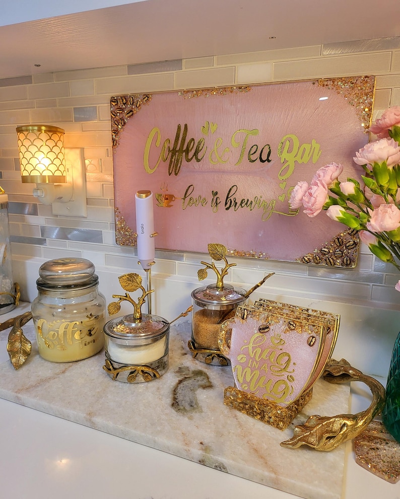 May include: A coffee and tea bar setup with a pink sign that reads "Coffee & Tea Bar" in gold lettering. The setup includes a jar, sugar bowl, and coasters, all with gold accents. A decorative lamp is visible.