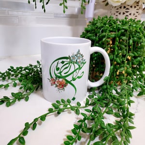 May include: White ceramic mug featuring green Arabic calligraphy and floral accents in shades of blue, orange, and pink. The mug has a curved handle and is surrounded by green leafy plants, suggesting a home decor theme.