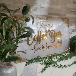 May include: A rectangular white sign with gold lettering that reads "Ford's Coffee Bar love is brewing." The sign has gold glitter accents on the edges and a coffee cup graphic. Decorative greenery and a vase are in the foreground.