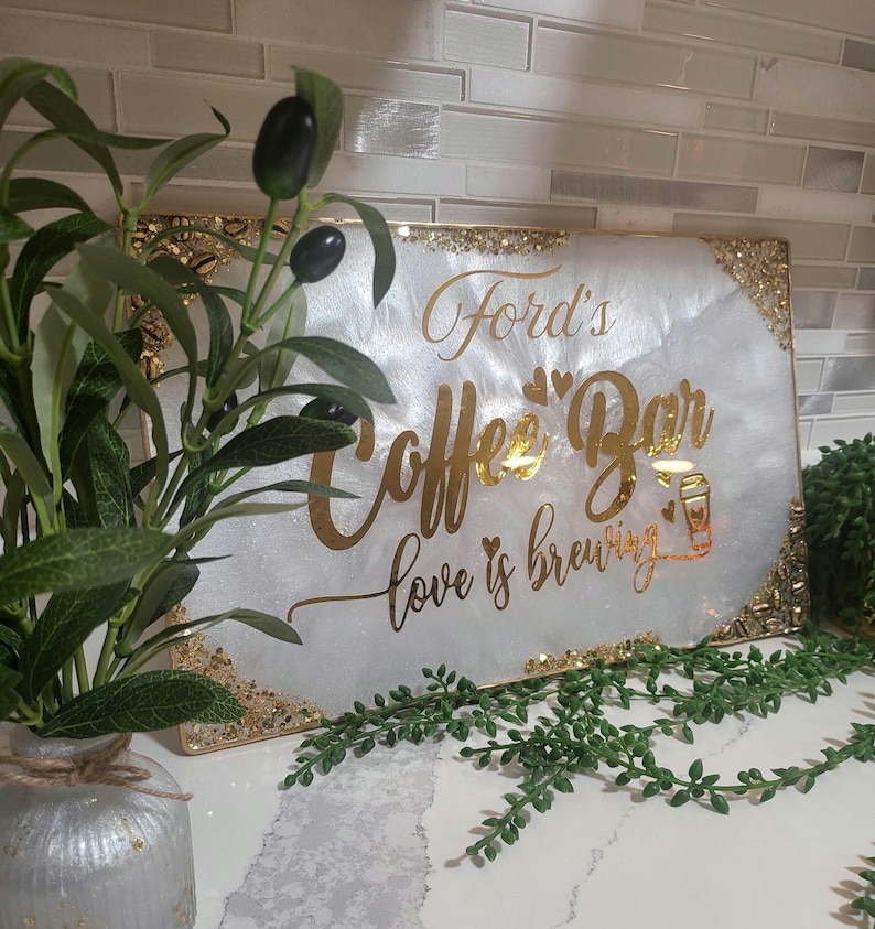 May include: A rectangular white sign with gold lettering that reads "Ford's Coffee Bar love is brewing." The sign has gold glitter accents on the edges and a coffee cup graphic. Decorative greenery and a vase are in the foreground.