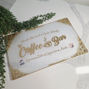May include: A rectangular coffee bar sign with a white background and gold glitter trim. The sign features the words "Coffee Bar" in gold script, along with coffee-related terms and phrases.