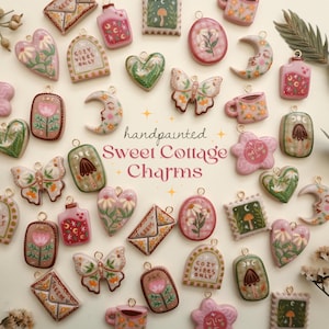 May include: Hand-painted Sweet Cottage charms in various shapes, including hearts, moons, butterflies, and flowers. The charms are in pastel colors with floral and nature-inspired designs. Some charms have the text "Cozy Vibes Only".
