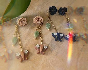 May include: Three pairs of dangle earrings with floral and butterfly charms. The earrings feature gold chains with green leaves and white flowers. The butterflies are in shades of brown, blue, and black.