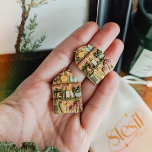 May include: A pair of handmade polymer clay earrings featuring a whimsical woodland scene. Each earring is shaped like a small arched doorway with a wooden frame, vines, and a variety of magical elements, including mushrooms, candles, books, and a crescent moon.