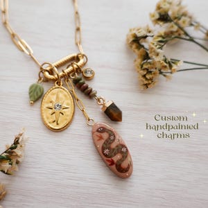 May include: Gold-toned necklace with multiple charms, including a starburst pendant, a green leaf, and a hand-painted charm with a snake design. The necklace also features a gemstone and the text "Custom handpainted charms".