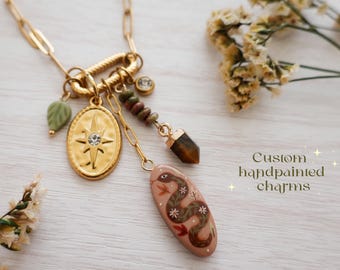 May include: Gold-toned necklace with multiple charms, including a starburst pendant, a green leaf, and a hand-painted charm with a snake design. The necklace also features a gemstone and the text "Custom handpainted charms".