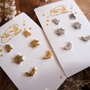 May include: Two sets of Halloween themed earrings on white cards. Each set includes a ghost, a spiderweb, and a bat. The earrings are made of gold and silver metal.