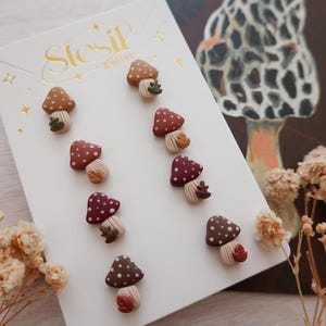 May include: A set of mushroom-shaped stud earrings in various colors, including brown, burgundy, and tan, with white polka dots. The earrings are displayed on a white card with the text "Stesil Jewelry".