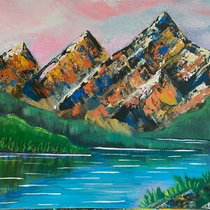 May include: An acrylic painting of a mountain landscape. The artwork depicts three large mountains with orange, blue, and white textured surfaces. A lake reflects the mountains and green trees under a pink and blue sky.