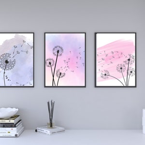 May include: Set of three framed art prints featuring dandelion illustrations. Each print has a black frame and showcases a dandelion design with a watercolor background in shades of purple, pink, and white. The artwork is displayed on a white surface.