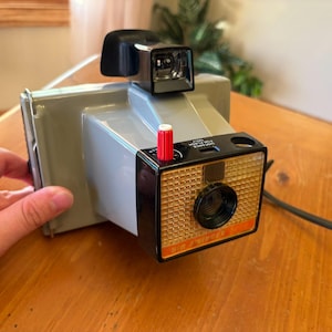 May include: A vintage Polaroid camera, featuring a gray body with a black and orange front panel. The camera has a red button and a black flash attachment. The lens is black, and the camera is held by a hand.
